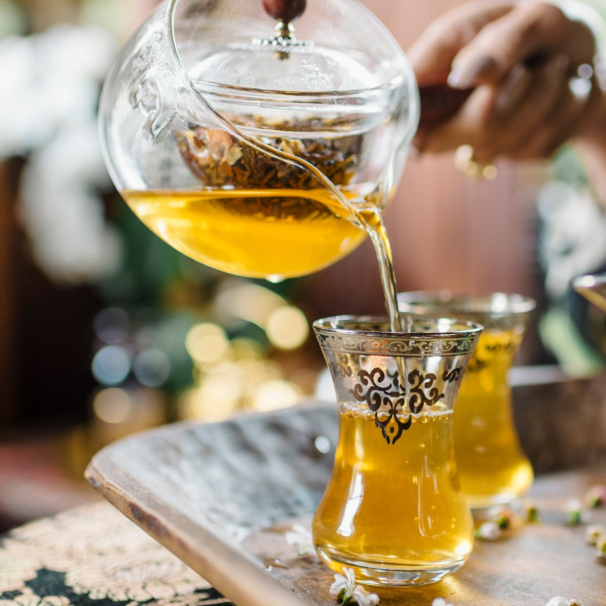 A hand pours yellow herbal tea—likely from the Oolong Tea Sampler Box—into ornate glass cups on a metal tray, with small white flowers scattered around.