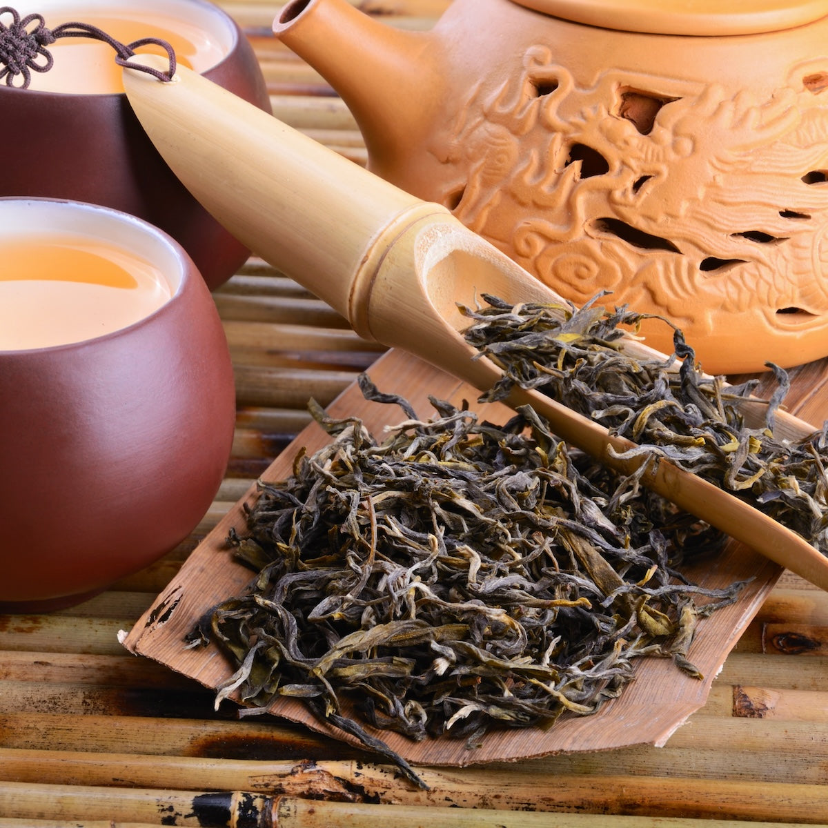 A close-up of a tea set featuring Bai Hao Super Fancy Formosa Oolong from Magic Hour on a wooden tray, accompanied by a bamboo tea scoop, two brown ceramic tea cups filled with this exquisite Taiwanese tea, and an intricately detailed clay teapot on a bamboo mat background. The scene provides a warm and traditional tea-drinking atmosphere.