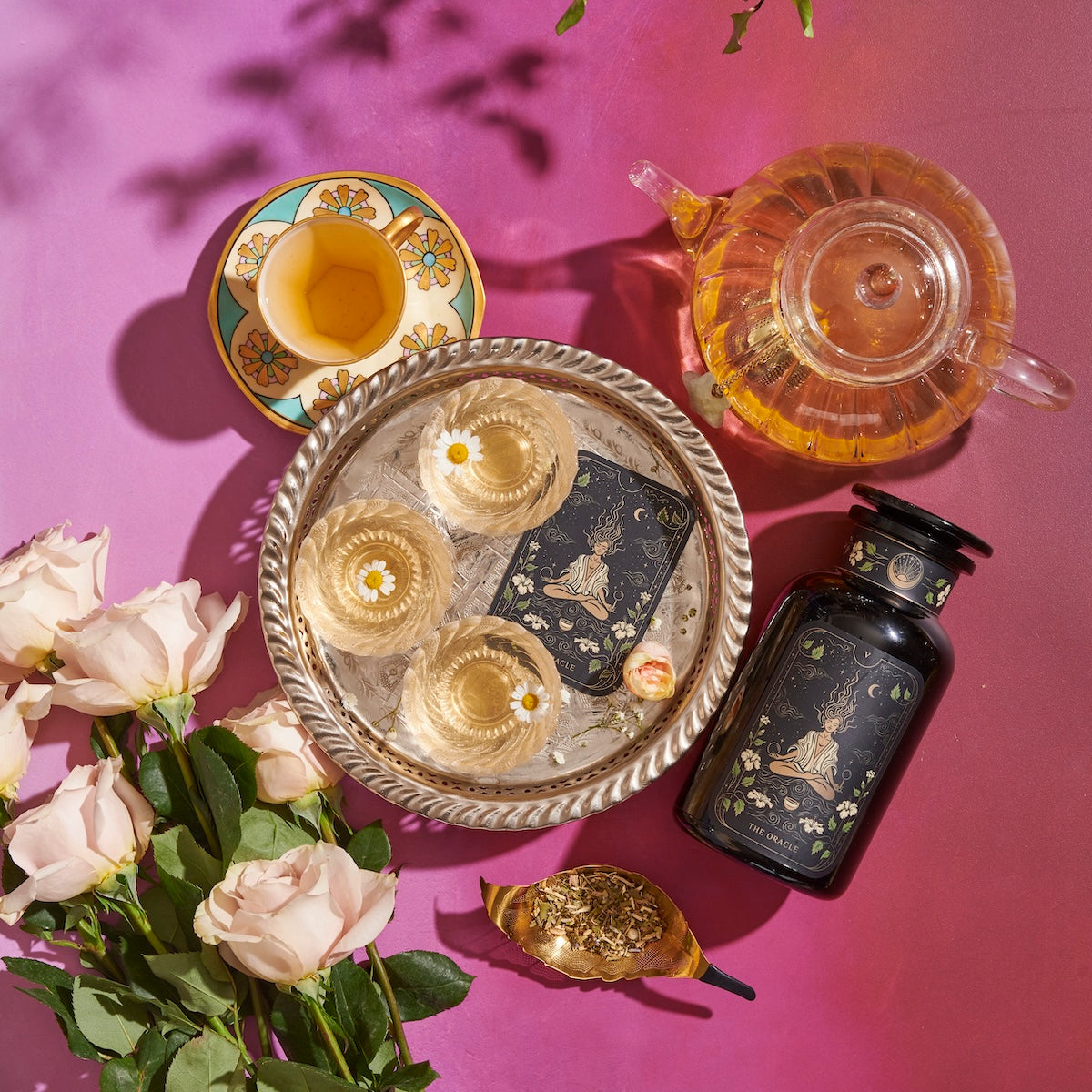 A tea setup with a teapot filled with wellness teas, floral-patterned teacups, and a matching floral tea tin on a vibrant pink and purple surface. Fresh white roses and a spoon containing loose tea leaves from the Monthly Magic First Sips Tea Subscription Box by Magic Hour are also visible for an elegant, serene ambiance.
