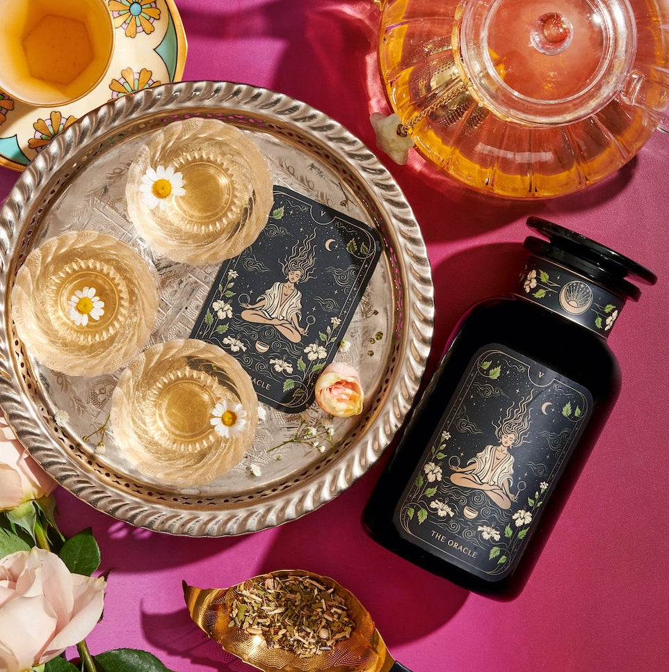 An elegant setup featuring a decorative tray with three glasses of champagne, a black bottle with an intricate label, Magic Hour's The Oracle: Lemon Lavender Tea for Supporting the Gut-Brain Axis in a clear teapot, a floral teacup, and a golden spoon with loose tea leaves, all arranged on a pink surface alongside Adaptogenic roots and white roses.