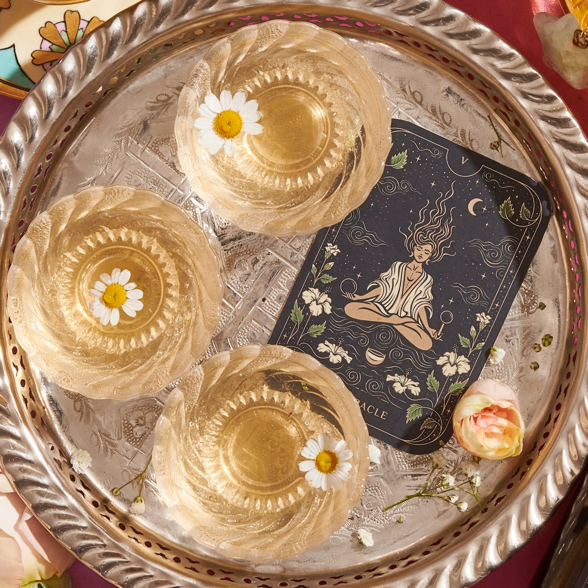 A decorative tray holds three round-shaped gel desserts with chamomile flowers on top. Beside them is an illustrated tarot card depicting a meditative figure amidst swirling lines, plants, and celestial elements. A small flower lies at the edge of the tray, hinting at a tranquil tarot journey enhanced by Magic Hour's Monthly Magic 3-Month Tea Subscription Box.