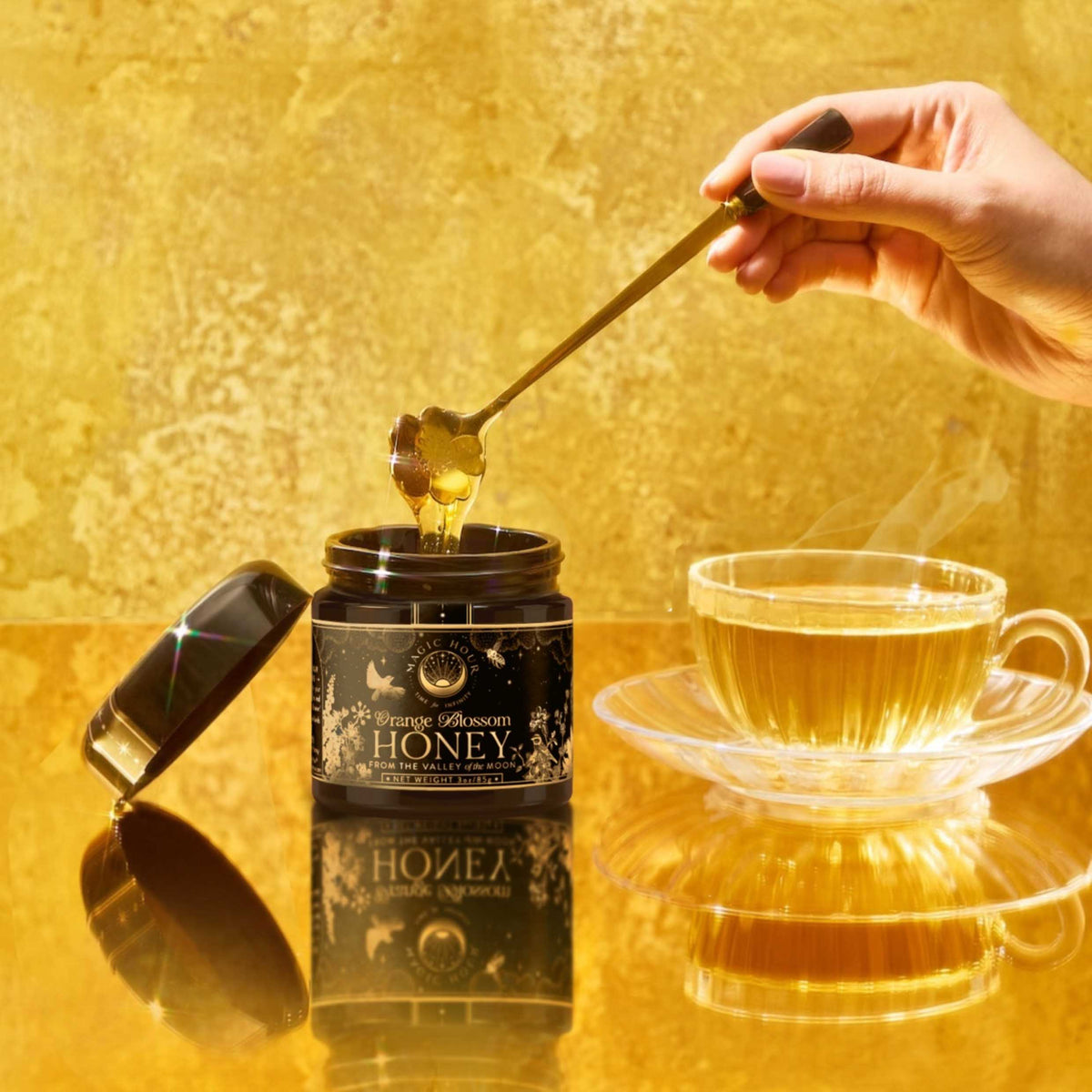 A hand holds a golden honey dipper over an open jar of Orange Blossom Honey: Raw Ojai Honey from the Valley of the Moon next to a cup of steaming tea.