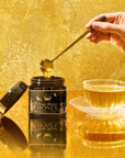 A hand holds a golden honey dipper over an open jar of Orange Blossom Honey: Raw Ojai Honey from the Valley of the Moon next to a cup of steaming tea.
