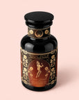 A dark glass jar labeled “Magic Hour: Oshun’s Nectar: Honeyed Herbal Tea for Joy & Radiance,” adorned with ornate gold floral designs and an illustration of a woman holding a flute. This golden honeyed herbal tea combines soothing rooibos honeybush tea with delicate botanicals, offering a truly enchanting experience.