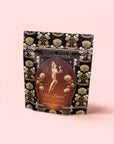 A floral-patterned bag from the Monthly Magic Tea Journey Subscription, featuring “She Rises: Oshun’s Nectar” honeyed herbal tea inspired by goddess archetypes, with an illustration of a woman adorned with flowers and gold accents on a peach background.