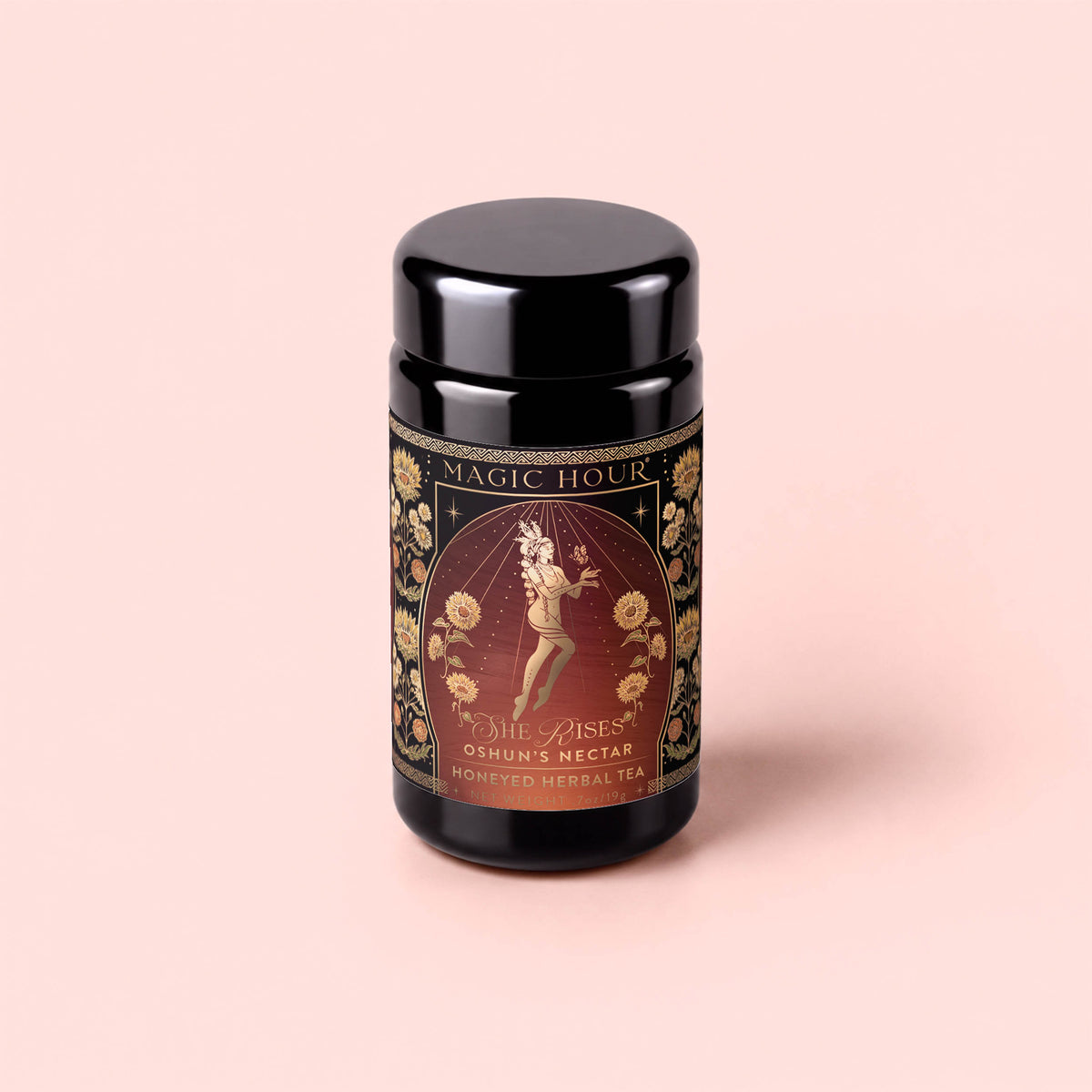 A black jar labeled “Magic Hour” with ornate gold floral designs and an illustration of a woman, featuring the product name “Oshun’s Nectar: Honeyed Herbal Tea for Joy &amp; Radiance.” The jar sits on a pale pink background.