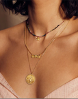 Close-up of a person wearing layered necklaces. The necklaces include a choker with colorful beads and a small pendant, a short chain with a gold bow pendant, and the Divine Mother Necklace featuring a round gold medallion depicting Our Lady of Guadalupe. The person is wearing a pink off-shoulder top.