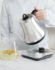 A person in a white jacket is performing a tea ceremony, carefully pouring hot water from the Magic Hour stainless steel gooseneck kettle into a glass pitcher containing loose tea. The setup is on a white marble countertop, with the Magic Hour kettle resting on its digital base featuring buttons and a small display screen.