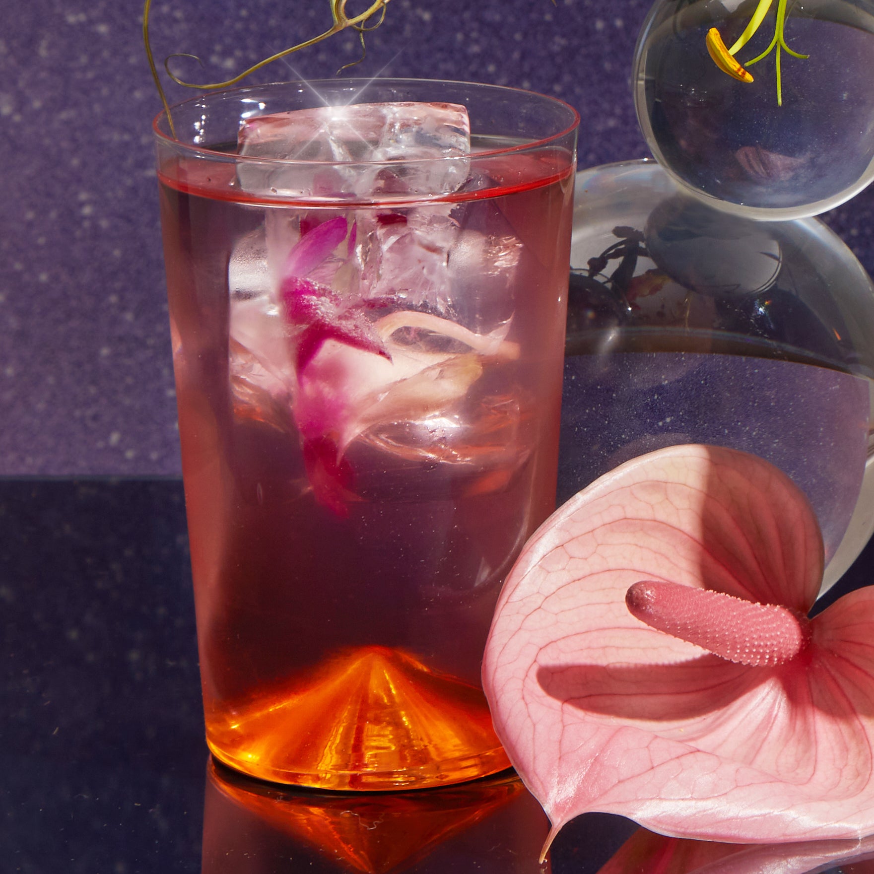 A clear glass of Cosmic Garden Iced Tea topped with a pink petal sits on a reflective surface beside pink decor and an anthurium, against a purple backdrop—the perfect way to enjoy the Cosmic Garden Iced Tea Sampler Set.