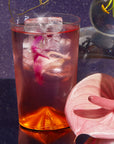 A clear glass of Cosmic Garden Iced Tea topped with a pink petal sits on a reflective surface beside pink decor and an anthurium, against a purple backdrop—the perfect way to enjoy the Cosmic Garden Iced Tea Sampler Set.