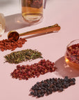 A pink surface displaying an assortment of dried herbs and berries in small piles, including goji berries, dried green leaves, rose petals, and dark berries. A gold measuring spoon and a clear cup with Passionfruit Green with Roses Iced Tea from Magic Hour are also present, along with a white rose in the background.