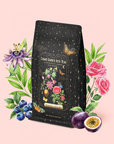 A black tea package labeled "Passionfruit Green with Roses Iced Tea" is surrounded by illustrated roses, blueberries, organic rose petals, passionfruit, green leaves, and golden sparkles—evoking the refreshing blend of Passionfruit green tea—against a pale pink background.