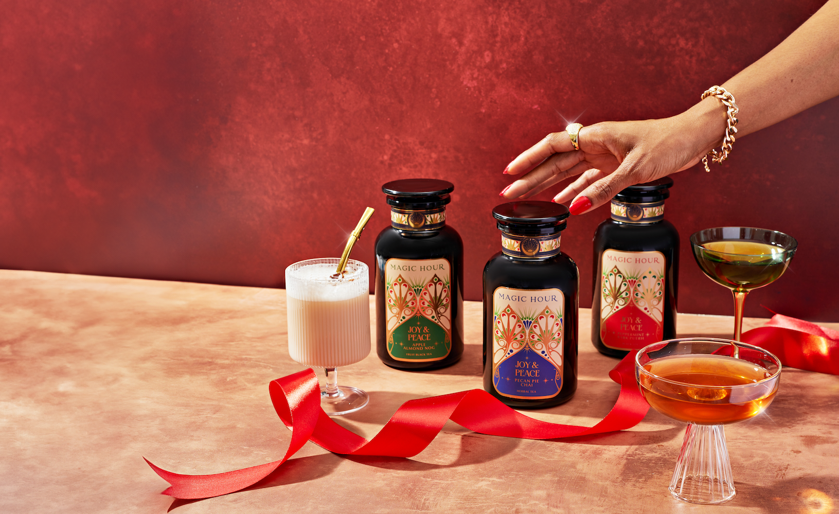 A hand reaches for one of three bottles labeled "Magic Hour" on a marble surface against a red backdrop. In front of the bottles are three different beverages in elegant glasses, accompanied by a red ribbon. The scene appears festive and elegant.