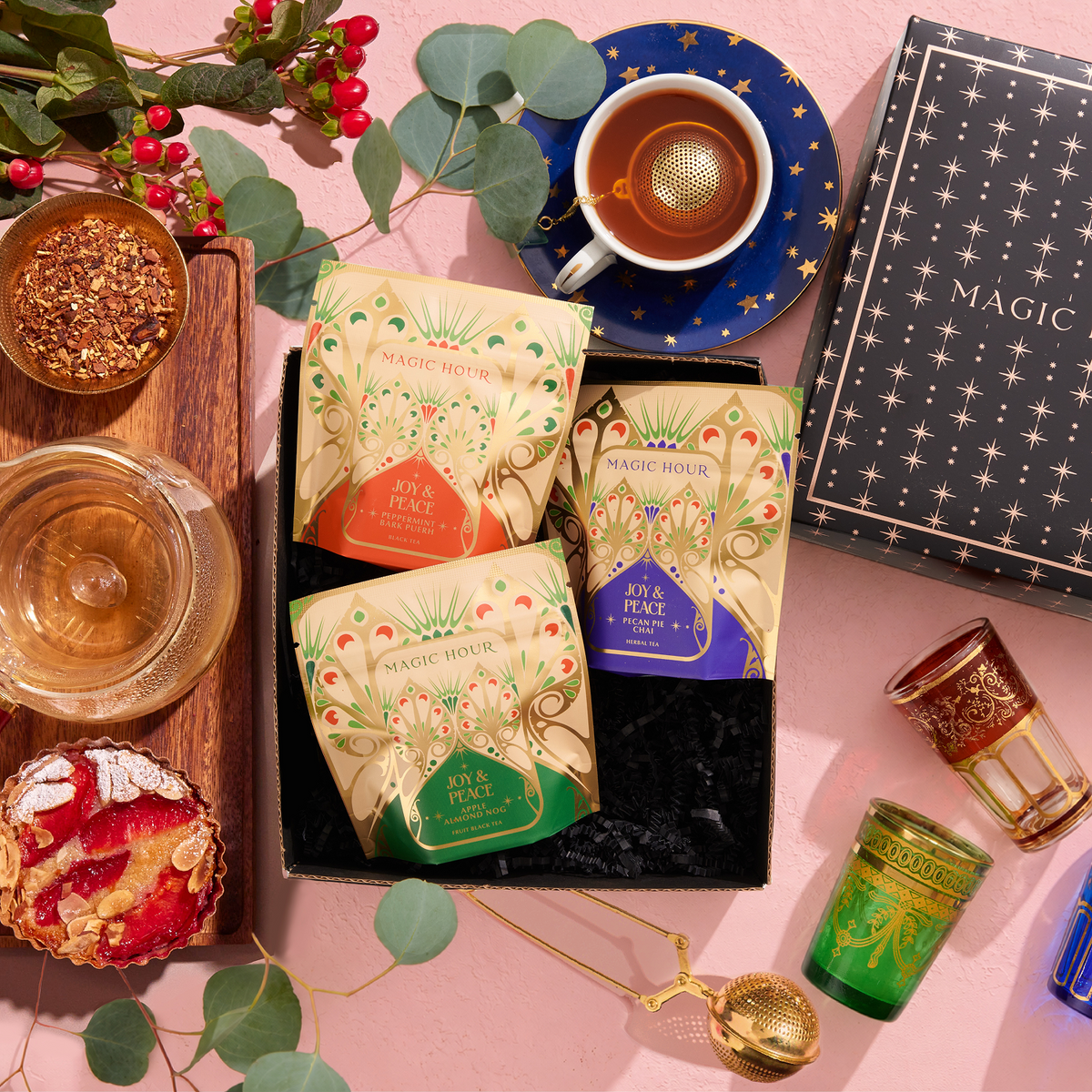 Peace & Joy Sampler Set featuring three tea pouches in a black box surrounded by a cup of tea, loose tea, a tart, a tea strainer, and two decorative glasses.