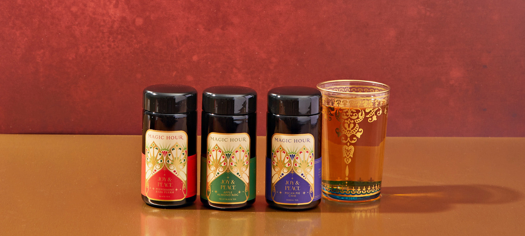 Three jars of Magic Hour tea, each with vibrant labels in red, green, and blue, are lined up next to a decorative glass filled with tea. The background is a rich reddish-brown, and the surface is a smooth gold color, creating a luxurious setting.