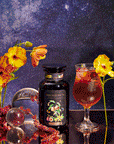 A glass of Peach Vanilla Black Iced Tea with Figs, a decorative tea tin, and yellow flowers sit on a mirrored surface under a starry sky. Glass spheres are scattered among the blooms for an elegant display.
