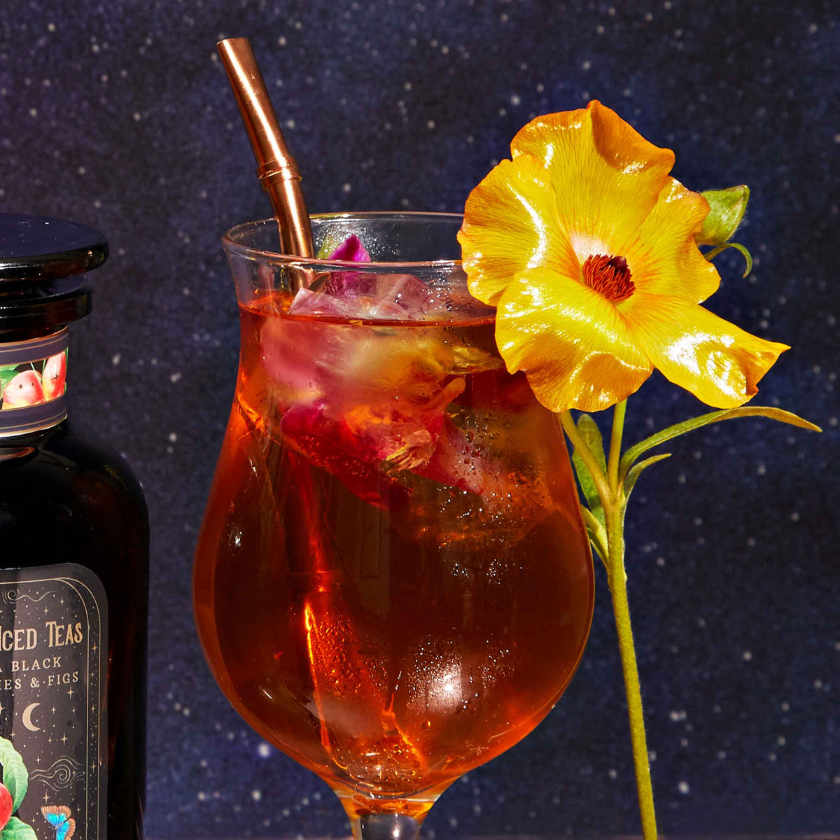A glass of iced tea with the Sips Tea Straw & Strainer Bombilla, garnished with a bright yellow flower, sits against a starry night background. A bottle labeled “Spiced Teas” is partially visible on the left, hinting at the rich flavors of loose tea within.