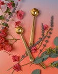 The Magic Hour Starter Kit by Magic Hour features two elegant gold measuring spoons placed among an array of vibrant flowers and leaves, all set against a gradient background that smoothly shifts from blue at the top to pink at the bottom. These spoons, evoking the essence of a tea ceremony, are artfully positioned to enhance the beauty of the floral display.