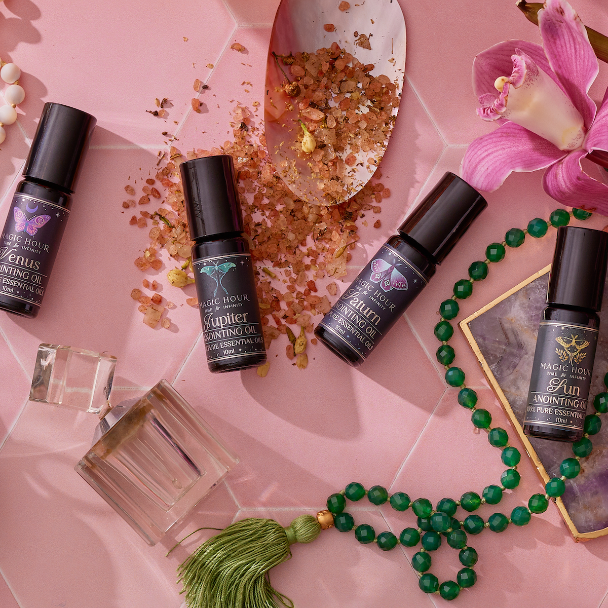 The Planetary Perfume Bundle (Save $26 with the Bundle!), crystals, pink bath salts, a green beaded tassel necklace, and a pink orchid on a geometric surface evoke emotional balance and tranquility.