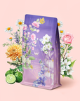 A pastel purple package of Perfumes of Provence: Lavender Bouquet Tea for Calm Moods and Beautiful Skin, adorned with floral and citrus illustrations, stands amid blooming flowers and bergamot fruit on a soft pink background, evoking the soothing essence of Provence.