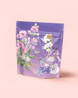 Perfumes of Provence: Lavender Bouquet Tea for Calm Moods and Beautiful Skin comes in a lavender resealable pouch adorned with pink, white, and purple flowers, green leaves, gold geometric accents, and delicate text on a soft pink background.