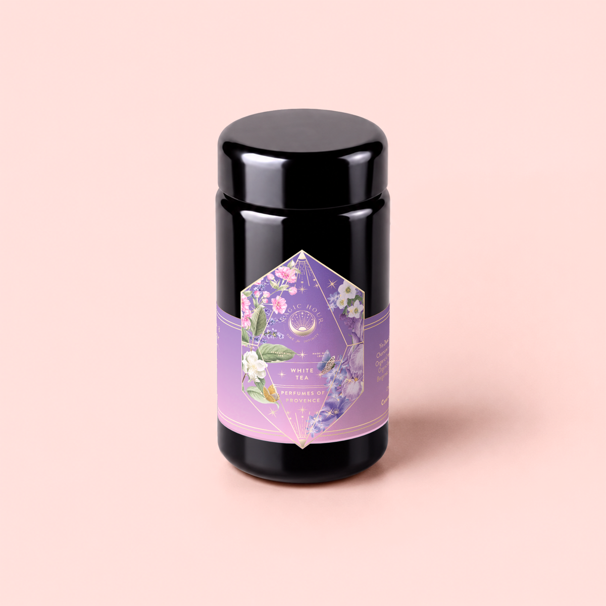 A black cylindrical tea canister with a purple floral label sits on a light pink background. The label features pastel flowers and green leaves, displaying the text “Perfumes of Provence: Lavender Bouquet Tea for Calm Moods and Beautiful Skin.” This calming blend evokes the gentle aroma of lavender in full bloom, offering an elegant and soothing tea experience.