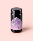 A black cylindrical tea canister with a purple floral label sits on a light pink background. The label features pastel flowers and green leaves, displaying the text “Perfumes of Provence: Lavender Bouquet Tea for Calm Moods and Beautiful Skin.” This calming blend evokes the gentle aroma of lavender in full bloom, offering an elegant and soothing tea experience.