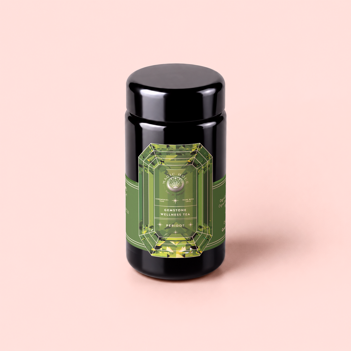 A black tea canister with a green, gem-shaped label reading “Peridot: Green Guayusa Oolong Mango-Peach Tea” stands on a light pink background, perfect for storing your favorite blends like guayusa or oolong tea.