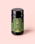 A black tea canister with a green, gem-shaped label reading “Peridot: Green Guayusa Oolong Mango-Peach Tea” stands on a light pink background, perfect for storing your favorite blends like guayusa or oolong tea.