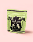 A green resealable pouch labeled "Piña Colada: Tropical Green Mocktail Tea for Sultry Moods & Radiant Skin," with palm trees and vintage swimmers on pale pink, offers refreshing organic green tea for a tropical mocktail experience.