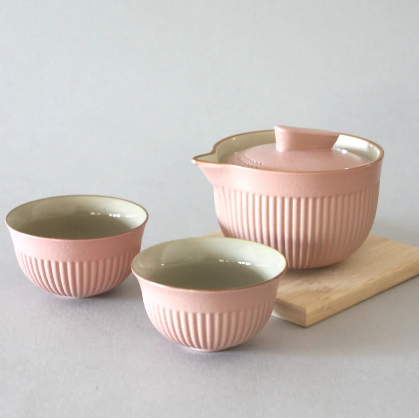 The Eastern Elm Pink Infuser Tea Set includes a high-quality ceramic teapot with a lid and two matching tea bowls, all in a pastel pink shade. Each item has vertical ribbed textures and is perfect for loose-leaf teas. This exquisite set is displayed on a light wooden tray against a plain, light gray background.