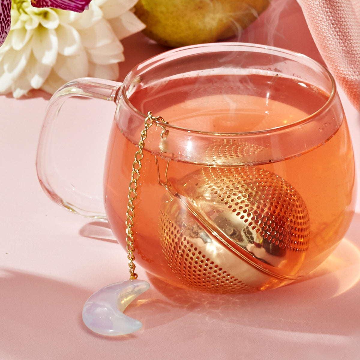 A clear glass teacup filled with reddish tea sits on a pink surface, paired with The Best of Fall Box. Inside, a gold mesh tea infuser attached to a chain with a crescent moon charm steams slightly, signaling hot tea. A white flower and part of a pear are visible in the background.