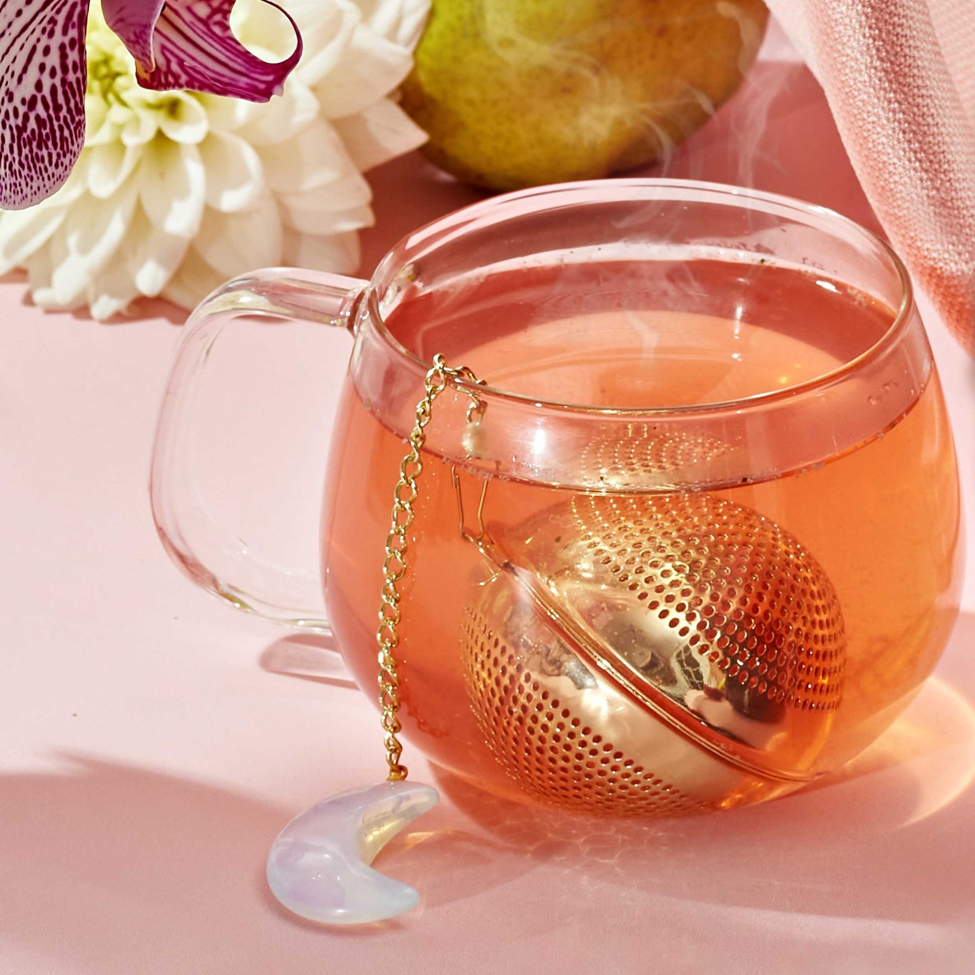 A glass mug of hot tea with a gold Opalite Moon Tea Strainer and a crescent moon charm sits on a pink surface, accompanied by a white flower, an orchid, and a pear in the background. Steam curls from the tea, enhancing the soothing ambiance of a spiritual tea ritual.
