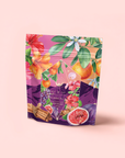A vibrant stand-up pouch with hibiscus flowers, oranges, figs, cinnamon sticks, and leaves on a pink-purple backdrop features Pink Moment Pixie - Hibiscus Tangerine Dream Herbal Tea—a caffeine-free fruity blend.