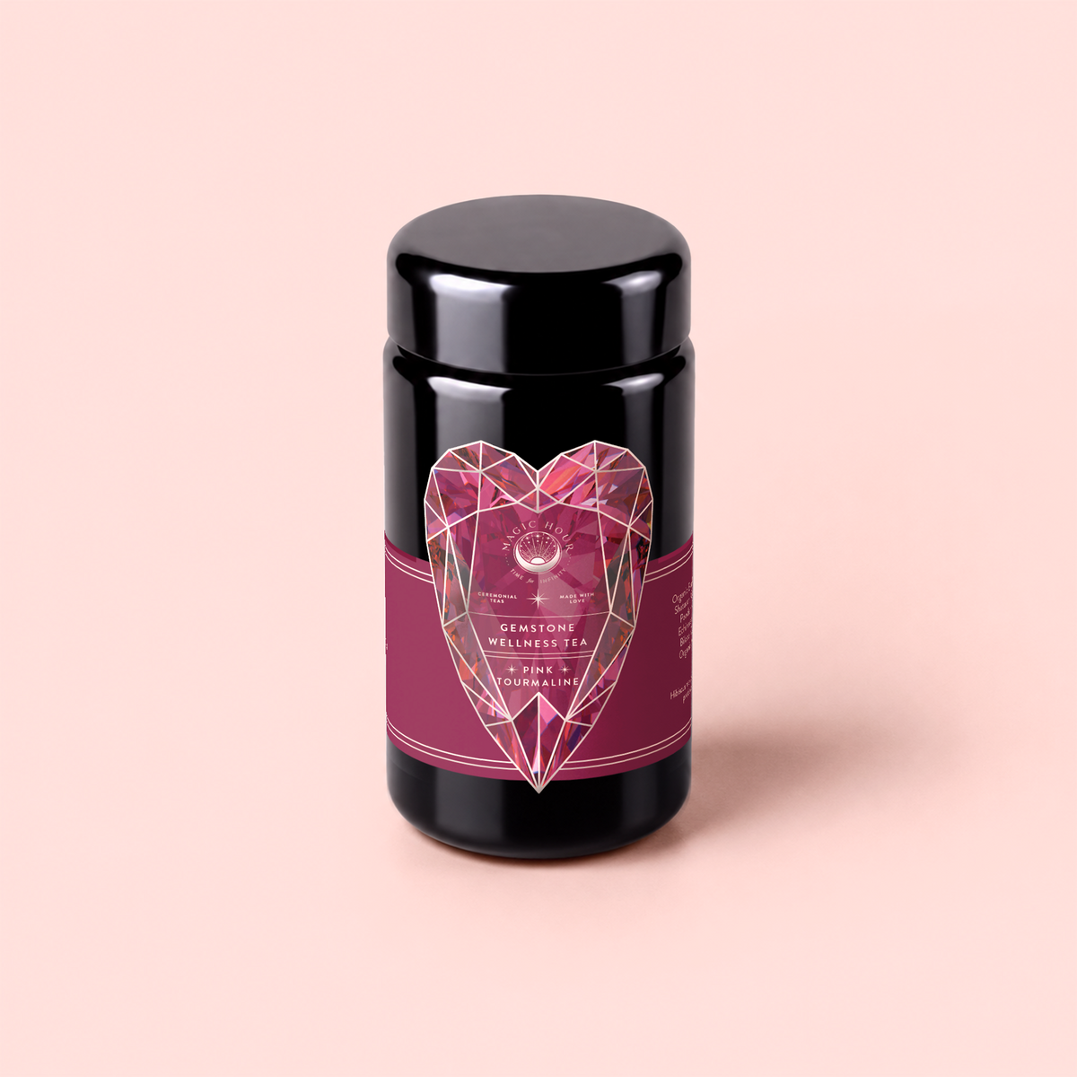 A canister of Pink Tourmaline: Organic Dragon fruit Hibiscus White Tea for Women’s Health, adorned with a pink label featuring a geometric heart design and the words “Gemstone Wellness Tea” and “Tourmaline,” sits on a light pink background. This thoughtfully crafted tea blend is designed to support heart and breast health while offering gentle immune support.