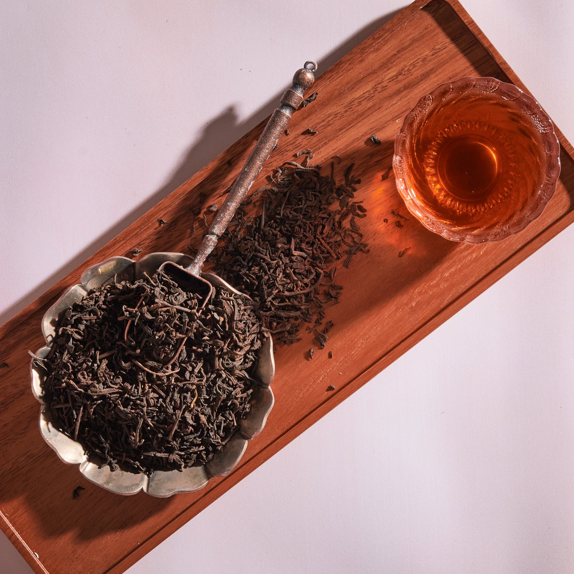 A wooden tray holds a bowl filled with loose leaf Sin Eraser™: Puerh Tea by Magic Hour and a spoon resting on top. Beside the bowl is a glass cup filled with brewed tea, casting a shadow on the tray. The scene suggests a tea preparation setup with warm, inviting aromas and rich health benefits.