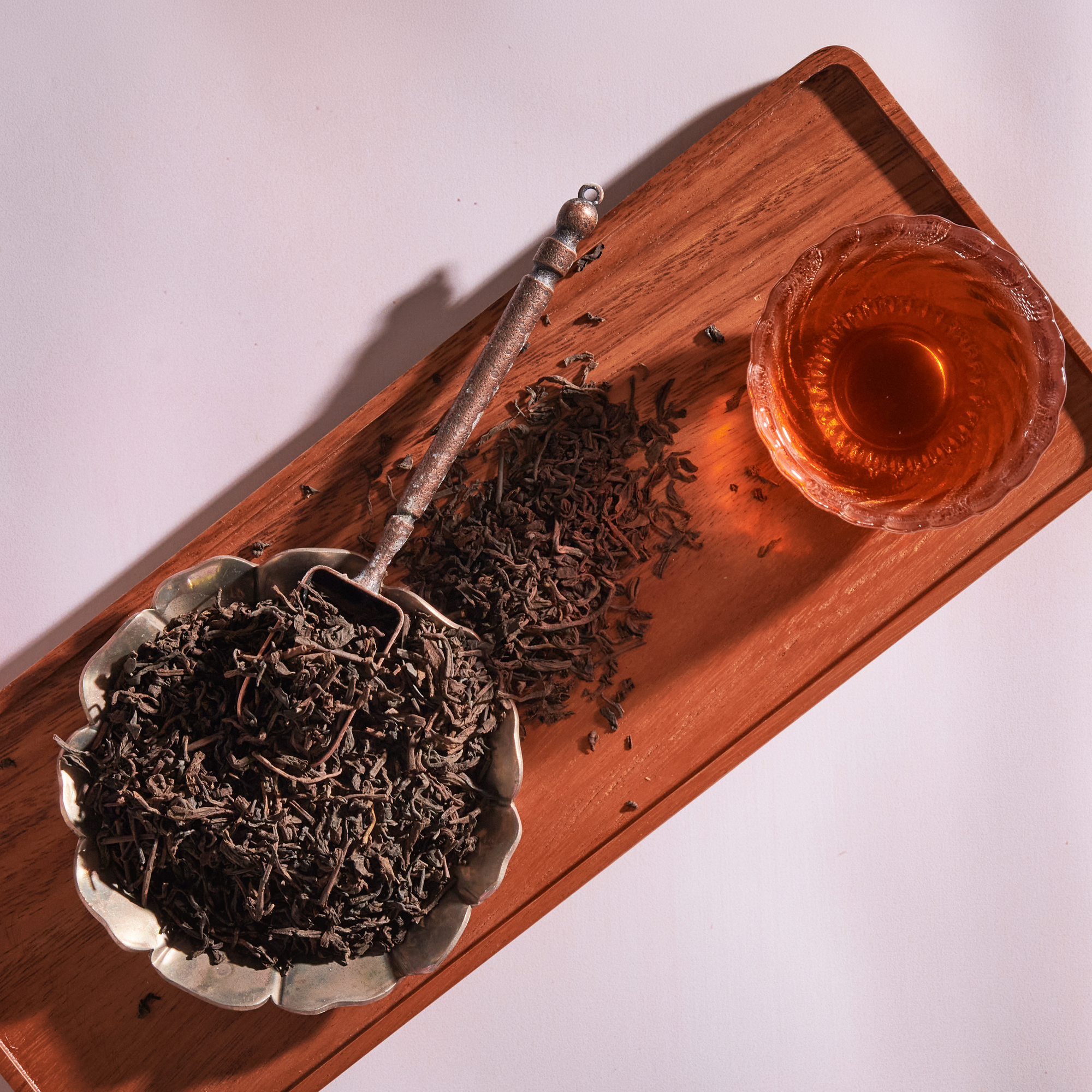Overhead view of loose Bohemian Breakfast Vanilla Puerh Black Tea for Digestion & Energy in a silver floral dish with a spoon on a wooden tray, beside a glass cup of brewed amber tea—a soothing coffee alternative.