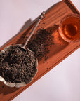 A wooden tray holds a bowl filled with loose leaf Sin Eraser™: Puerh Tea by Magic Hour and a spoon resting on top. Beside the bowl is a glass cup filled with brewed tea, casting a shadow on the tray. The scene suggests a tea preparation setup with warm, inviting aromas and rich health benefits.