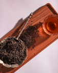 Overhead view of loose Bohemian Breakfast Vanilla Puerh Black Tea for Digestion & Energy in a silver floral dish with a spoon on a wooden tray, beside a glass cup of brewed amber tea—a soothing coffee alternative.