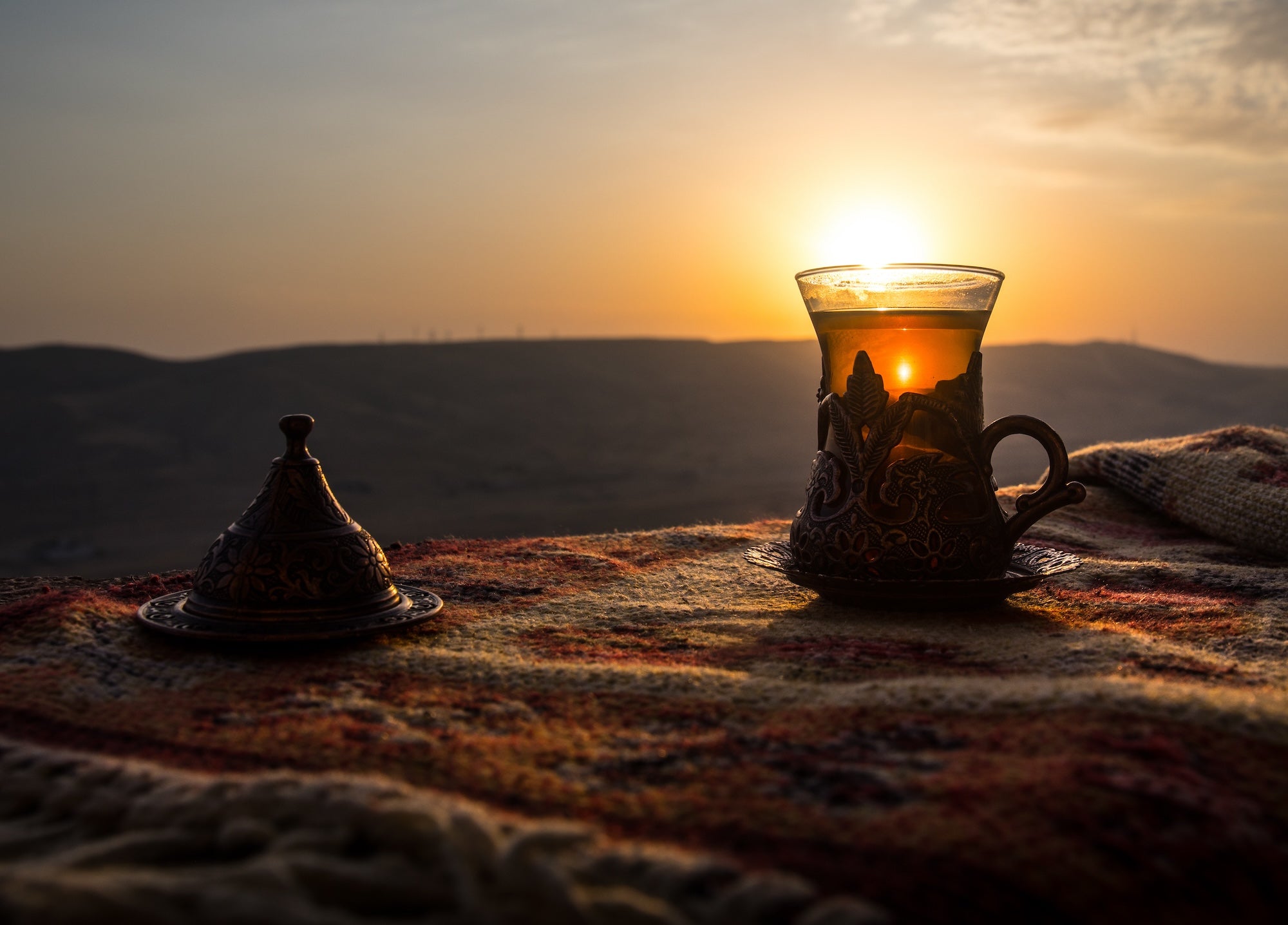 A decorative glass of tea and a small covered dish rest on a colorful woven cloth against a backdrop of rolling hills at sunset. The sun's rays create a warm, inviting glow, highlighting the serene and peaceful ambiance.