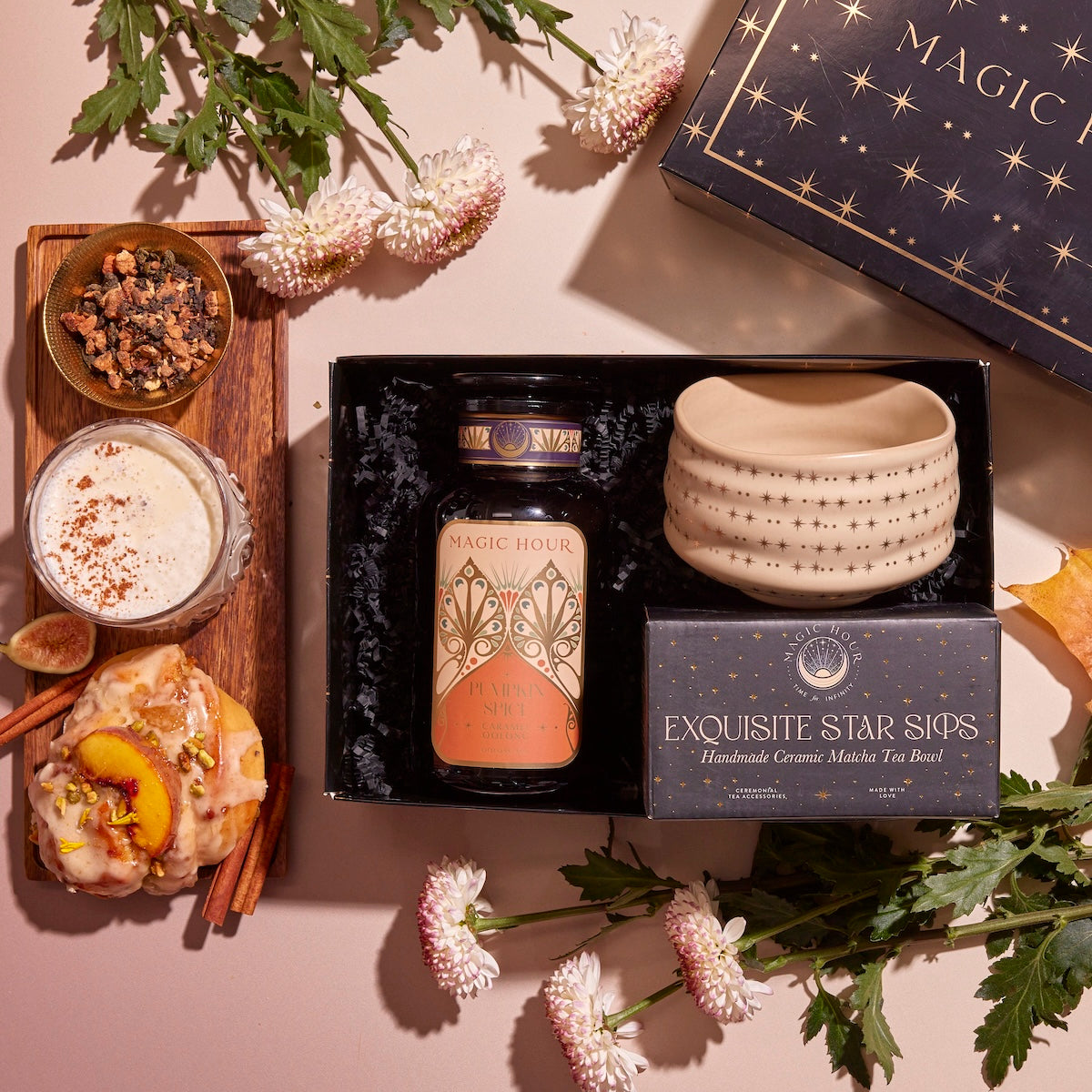 A "Pumpkin Lovers Tea & Accessory Gift Set" is elegantly displayed on a pink surface, featuring a box labeled "Magic Hour," a tin of pumpkin spice tea, a ceremonial tea bowl, and loose tea herbs. Accompanying the set are a latte with cinnamon and peach tart, surrounded by white flowers and green leaves to enhance the presentation.