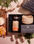 A "Pumpkin Lovers Tea & Accessory Gift Set" is elegantly displayed on a pink surface, featuring a box labeled "Magic Hour," a tin of pumpkin spice tea, a ceremonial tea bowl, and loose tea herbs. Accompanying the set are a latte with cinnamon and peach tart, surrounded by white flowers and green leaves to enhance the presentation.