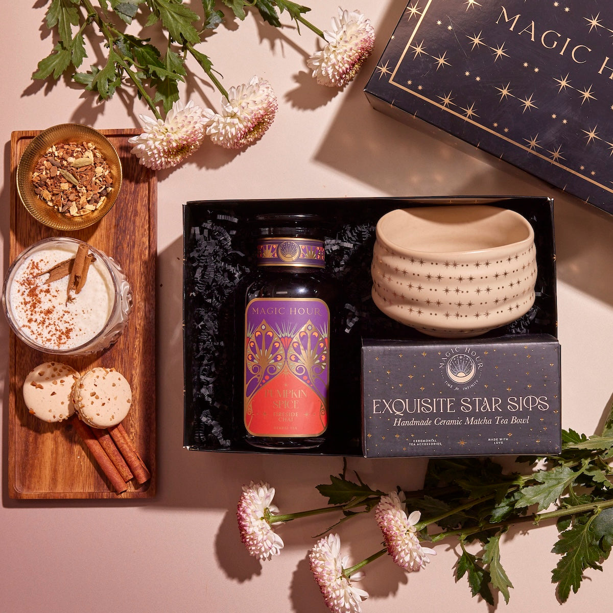 The Pumpkin Lovers Tea &amp; Accessory Gift Set includes a tea canister, a ceramic ceremonial tea bowl, and an &quot;Exquisite Star Sips&quot; package. Alongside it, you&#39;ll find a frothy pumpkin spice tea accompanied by cinnamon sticks, cookies, and white flowers beautifully displayed on a wooden board.