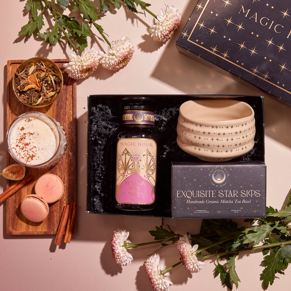 The Pumpkin Lovers Tea & Accessory Gift Set includes a jar labeled "Magic Hour," a box of "Exquisite Star Sips," a luxurious ceremonial tea bowl, and a cup of pumpkin spice tea. It comes paired with macarons and cinnamon sticks, all beautifully arranged on a wooden tray with decorative flowers elegantly surrounding these delightful items.