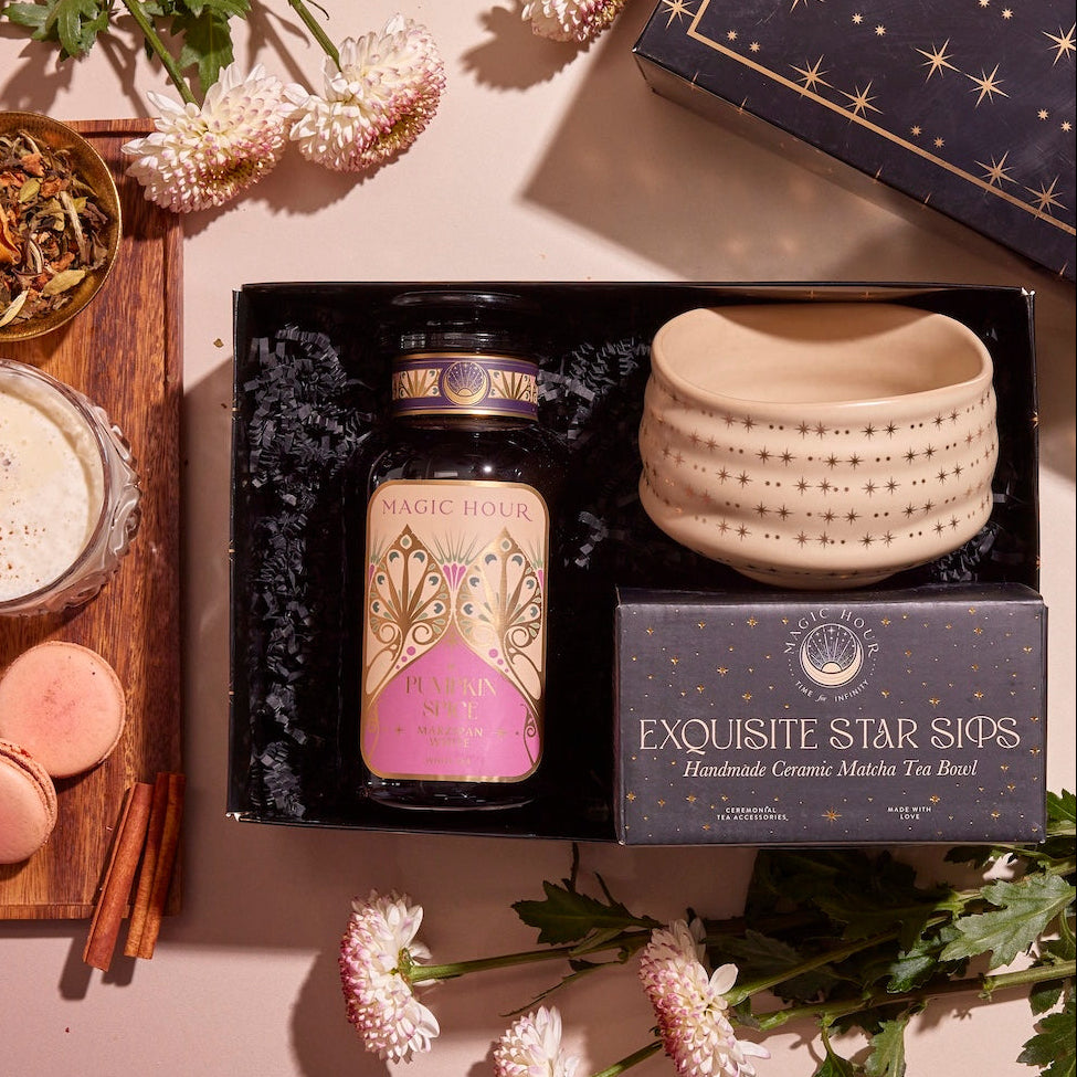 The Pumpkin Lovers Tea & Accessory Gift Set includes a jar labeled "Magic Hour," a box of "Exquisite Star Sips," a luxurious ceremonial tea bowl, and a cup of pumpkin spice tea. It comes paired with macarons and cinnamon sticks, all beautifully arranged on a wooden tray with decorative flowers elegantly surrounding these delightful items.