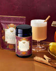 Pumpkin Spice Matcha packaging, a jar, and a latte in a glass sit on a table surrounded by a pumpkin, pears, and cinnamon sticks against a warm reddish-brown backdrop—a cozy scene that beautifully embodies the essence of autumn botanicals.