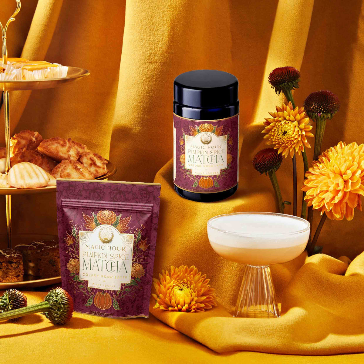 Pumpkin Spice Matcha jars and a drink are displayed on gold fabric surrounded by autumn botanicals, vibrant flowers, and pastries, creating a cozy fall atmosphere.