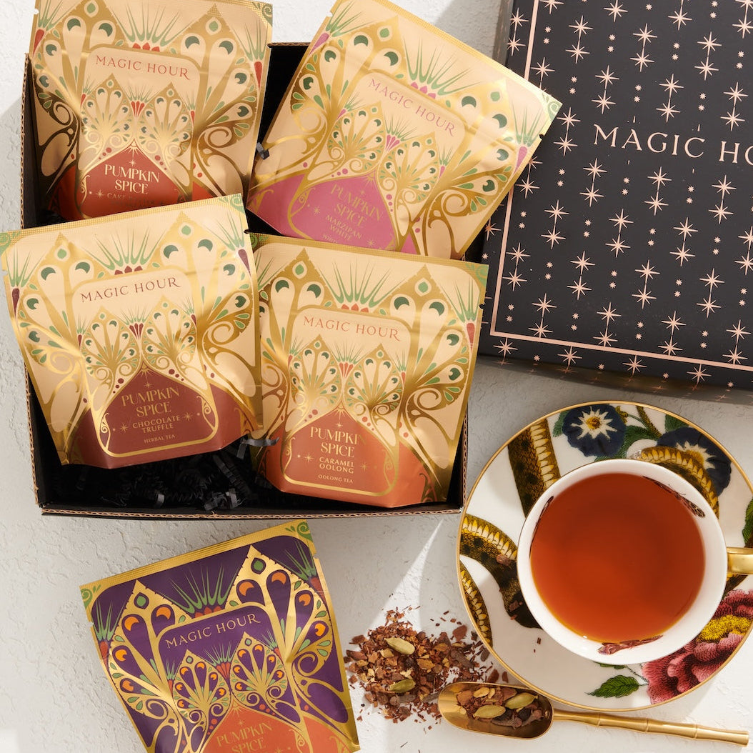 A stylishly arranged photo featuring a box of "Pumpkin Spice Abundance Sampler Bundle" tea packets in festive, decorative packaging. Next to the box is a teacup filled with tea, adorned with floral designs. Some tea leaves are scattered by the cup, adding a charming touch reminiscent of autumn's warmth and imaginative flavors.