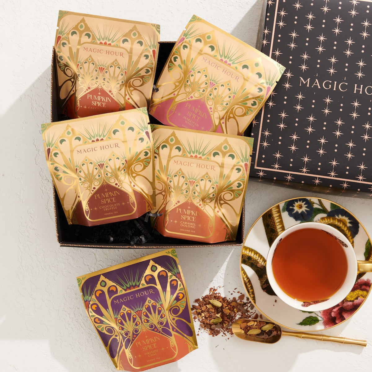 A stylishly arranged photo featuring a box of "Pumpkin Spice Abundance Sampler Bundle" tea packets in festive, decorative packaging. Next to the box is a teacup filled with tea, adorned with floral designs. Some tea leaves are scattered by the cup, adding a charming touch reminiscent of autumn's warmth and imaginative flavors.