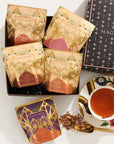 A stylishly arranged photo featuring a box of "Pumpkin Spice Abundance Sampler Bundle" tea packets in festive, decorative packaging. Next to the box is a teacup filled with tea, adorned with floral designs. Some tea leaves are scattered by the cup, adding a charming touch reminiscent of autumn's warmth and imaginative flavors.